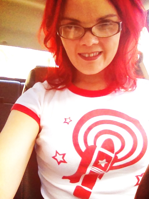 JoEllen sitting in a car wearing brown glasses and a white t-shirt with red rings on the sleeves and neck and a red vibrator design on the front